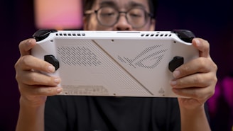 A person holds a white handheld gaming device with black controls, featuring a futuristic design on its back. The person is focused on the device with a blurred background creating a sense of depth.