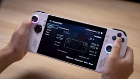 A person is holding a handheld gaming console that features a screen displaying a menu for gamepad mode. The console has control sticks and buttons located on both sides of the screen. The background appears to be a wooden surface.