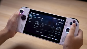 A person is holding a handheld gaming console that features a screen displaying a menu for gamepad mode. The console has control sticks and buttons located on both sides of the screen. The background appears to be a wooden surface.
