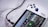 A handheld gaming console with a white body and colorful buttons features a screen displaying various game icons. The device is connected to a power source via a black cable and rests on a marble surface. The interface shows games like Apex and Street Fighter.