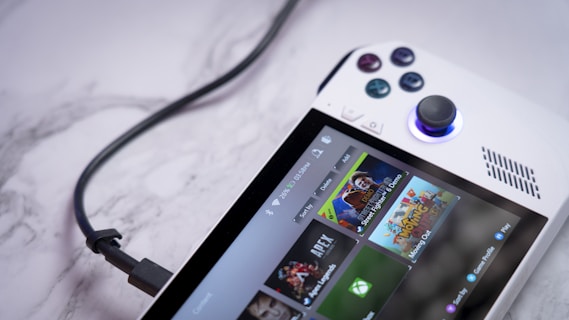 A handheld gaming console with a white body and colorful buttons features a screen displaying various game icons. The device is connected to a power source via a black cable and rests on a marble surface. The interface shows games like Apex and Street Fighter.