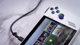 A handheld gaming console with a white body and colorful buttons features a screen displaying various game icons. The device is connected to a power source via a black cable and rests on a marble surface. The interface shows games like Apex and Street Fighter.