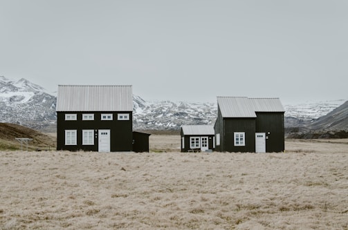 Three dark-colored houses with white roofs are situated in a vast, open landscape with fields in the foreground. Snow-capped mountains stretch across the horizon under an overcast sky, creating a striking contrast with the simple architecture.