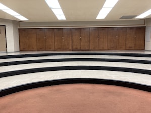 A series of wooden lockers positioned against a wall in a room with a curved, tiered floor. The tiers are made of black and white tiles, and there's a brown carpeted area in the foreground. The ceiling features bright fluorescent lights.