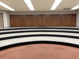 A series of wooden lockers positioned against a wall in a room with a curved, tiered floor. The tiers are made of black and white tiles, and there's a brown carpeted area in the foreground. The ceiling features bright fluorescent lights.