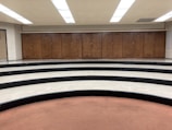 A series of wooden lockers positioned against a wall in a room with a curved, tiered floor. The tiers are made of black and white tiles, and there's a brown carpeted area in the foreground. The ceiling features bright fluorescent lights.