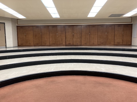A series of wooden lockers positioned against a wall in a room with a curved, tiered floor. The tiers are made of black and white tiles, and there's a brown carpeted area in the foreground. The ceiling features bright fluorescent lights.