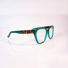 A pair of eyeglasses with a distinctive design, featuring green transparent frames with brown tortoiseshell accents on the temples. The glasses are set against a plain, soft-focus background, highlighting their stylish and modern appearance.