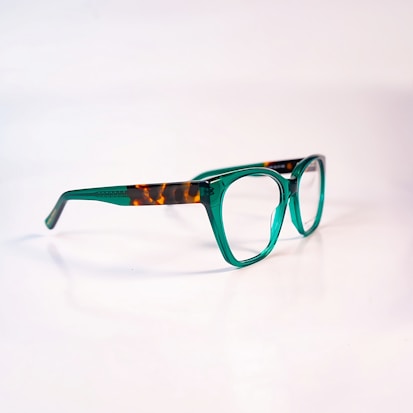 A pair of eyeglasses with a distinctive design, featuring green transparent frames with brown tortoiseshell accents on the temples. The glasses are set against a plain, soft-focus background, highlighting their stylish and modern appearance.