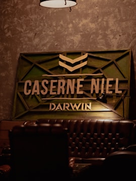 The image features a dimly lit interior with an industrial design. A vintage leather couch is partially visible in the foreground. On the wall, there is a wooden sign with green accents and the words CASERNE NIEL and DARWIN in bold letters. A lamp hangs from the ceiling above the scene.