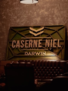 The image features a dimly lit interior with an industrial design. A vintage leather couch is partially visible in the foreground. On the wall, there is a wooden sign with green accents and the words CASERNE NIEL and DARWIN in bold letters. A lamp hangs from the ceiling above the scene.