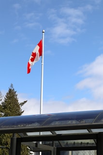 A Canadian flag is waving atop a tall flagpole against a blue sky with scattered clouds. Beneath the flag, a glass or transparent structure is partially visible along with some treetops.