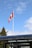 A Canadian flag is waving atop a tall flagpole against a blue sky with scattered clouds. Beneath the flag, a glass or transparent structure is partially visible along with some treetops.