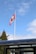 A Canadian flag is waving atop a tall flagpole against a blue sky with scattered clouds. Beneath the flag, a glass or transparent structure is partially visible along with some treetops.