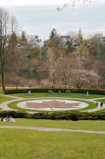 A landscaped park area features a large circular design with a leaf pattern made from earth and grass. Surrounding the circle is a well-maintained lawn with pathways that curve gently around the design. A few people are walking and others are seated on the grass. The background consists of a serene lake lined with trees, and beyond that, charming residential houses are nestled among more trees.