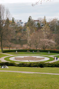 A landscaped park area features a large circular design with a leaf pattern made from earth and grass. Surrounding the circle is a well-maintained lawn with pathways that curve gently around the design. A few people are walking and others are seated on the grass. The background consists of a serene lake lined with trees, and beyond that, charming residential houses are nestled among more trees.