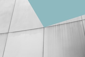 A minimalist architectural structure with large, smooth panels intersecting at sharp angles. The surface has an industrial look with subtle lines and a light shadow cast. The top portion of the image features a large triangular section of solid, muted teal color.