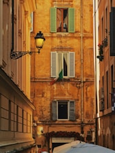 A narrow alleyway framed by historically styled buildings with a rustic ochre facade featuring three shuttered windows. The Italian flag is displayed prominently under one window. An ornate street lamp is mounted on the left building. Below, a sign for a restaurant named 'Achille Al Pantheon' and several umbrellas or canopies are visible.