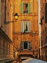 A narrow alleyway framed by historically styled buildings with a rustic ochre facade featuring three shuttered windows. The Italian flag is displayed prominently under one window. An ornate street lamp is mounted on the left building. Below, a sign for a restaurant named 'Achille Al Pantheon' and several umbrellas or canopies are visible.