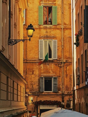 A narrow alleyway framed by historically styled buildings with a rustic ochre facade featuring three shuttered windows. The Italian flag is displayed prominently under one window. An ornate street lamp is mounted on the left building. Below, a sign for a restaurant named 'Achille Al Pantheon' and several umbrellas or canopies are visible.