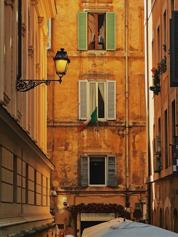 A narrow alleyway framed by historically styled buildings with a rustic ochre facade featuring three shuttered windows. The Italian flag is displayed prominently under one window. An ornate street lamp is mounted on the left building. Below, a sign for a restaurant named 'Achille Al Pantheon' and several umbrellas or canopies are visible.