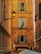 A narrow alleyway framed by historically styled buildings with a rustic ochre facade featuring three shuttered windows. The Italian flag is displayed prominently under one window. An ornate street lamp is mounted on the left building. Below, a sign for a restaurant named 'Achille Al Pantheon' and several umbrellas or canopies are visible.