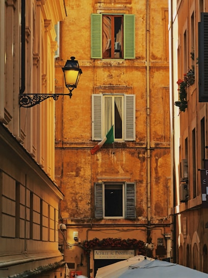 A narrow alleyway framed by historically styled buildings with a rustic ochre facade featuring three shuttered windows. The Italian flag is displayed prominently under one window. An ornate street lamp is mounted on the left building. Below, a sign for a restaurant named 'Achille Al Pantheon' and several umbrellas or canopies are visible.
