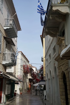 A narrow, picturesque alleyway with buildings on both sides, adorned with decorative balconies and vibrant red flowers. Greek flags are prominently displayed. Outdoor furniture lines the street, suggesting a cafe setting with a serene and charming Mediterranean atmosphere.