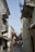 A narrow, picturesque alleyway with buildings on both sides, adorned with decorative balconies and vibrant red flowers. Greek flags are prominently displayed. Outdoor furniture lines the street, suggesting a cafe setting with a serene and charming Mediterranean atmosphere.