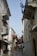 A narrow, picturesque alleyway with buildings on both sides, adorned with decorative balconies and vibrant red flowers. Greek flags are prominently displayed. Outdoor furniture lines the street, suggesting a cafe setting with a serene and charming Mediterranean atmosphere.