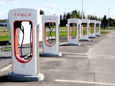 A row of Tesla electric car charging stations stands in a parking lot with clear skies in the background. Each station is white with red accents and the Tesla logo prominently displayed. The landscape around includes green grass and trees, with a building visible in the distance.