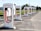 A row of Tesla electric car charging stations stands in a parking lot with clear skies in the background. Each station is white with red accents and the Tesla logo prominently displayed. The landscape around includes green grass and trees, with a building visible in the distance.