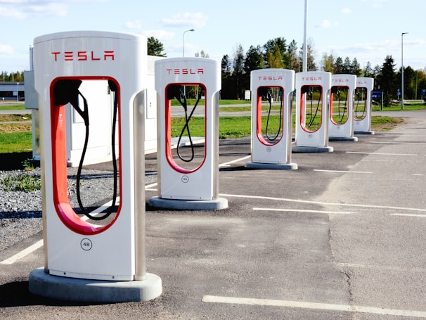 A row of Tesla electric car charging stations stands in a parking lot with clear skies in the background. Each station is white with red accents and the Tesla logo prominently displayed. The landscape around includes green grass and trees, with a building visible in the distance.