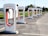 A row of Tesla electric car charging stations stands in a parking lot with clear skies in the background. Each station is white with red accents and the Tesla logo prominently displayed. The landscape around includes green grass and trees, with a building visible in the distance.