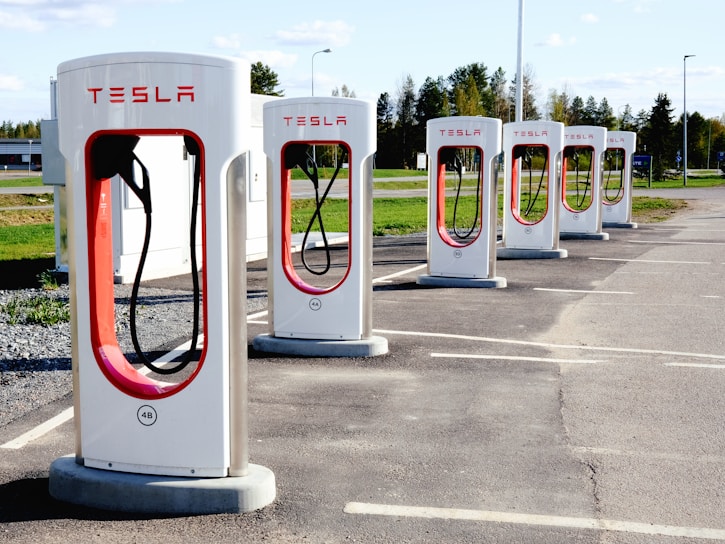A row of Tesla electric car charging stations stands in a parking lot with clear skies in the background. Each station is white with red accents and the Tesla logo prominently displayed. The landscape around includes green grass and trees, with a building visible in the distance.