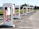 A row of Tesla electric car charging stations stands in a parking lot with clear skies in the background. Each station is white with red accents and the Tesla logo prominently displayed. The landscape around includes green grass and trees, with a building visible in the distance.