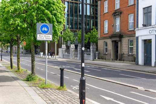A quiet urban street with a designated cycle track is lined with green leafy trees. There are modern and older brick buildings on either side. A blue sign indicates a cycle track in both English and Irish. The street appears clean and well-maintained, and there are a few people in the background.