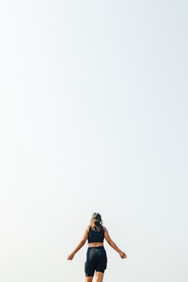 A person is viewed from behind, standing with arms slightly extended in a spacious outdoor setting. The bright, clear sky dominates the background, contributing to a sense of openness and tranquility.