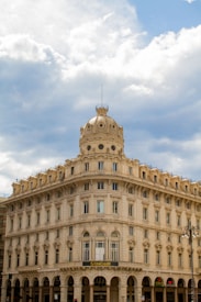 An ornate, historic building with multiple arched windows and a decorative dome on top sits beneath a partly cloudy sky. The structure is predominantly made of light-colored stone and features classical architectural elements. A UniCredit bank sign is visible above the entrance, indicating commercial use.