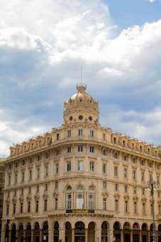 An ornate, historic building with multiple arched windows and a decorative dome on top sits beneath a partly cloudy sky. The structure is predominantly made of light-colored stone and features classical architectural elements. A UniCredit bank sign is visible above the entrance, indicating commercial use.