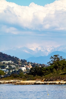 A coastal scene featuring a tranquil body of water in the foreground, lined by rocky shores. The mid-ground displays lush greenery and a cluster of houses on a hillside, with varied architectural styles. The background presents majestic, snow-capped mountains under a sky filled with large, billowing clouds.