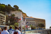 A group of people are walking towards a coastal hotel with large signage visible. The building is situated near a cliff adorned with greenery and other structures. Various flags, including one of France, are displayed prominently. The sky is clear and blue.