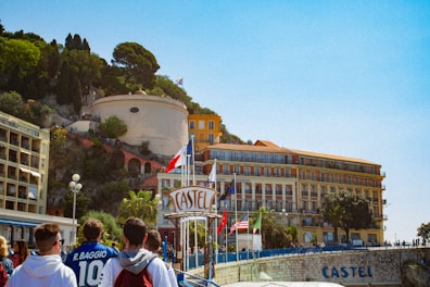 A group of people are walking towards a coastal hotel with large signage visible. The building is situated near a cliff adorned with greenery and other structures. Various flags, including one of France, are displayed prominently. The sky is clear and blue.