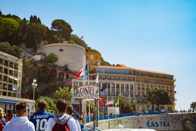 A group of people are walking towards a coastal hotel with large signage visible. The building is situated near a cliff adorned with greenery and other structures. Various flags, including one of France, are displayed prominently. The sky is clear and blue.
