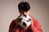 A person with dark hair partially obscured is holding a Telstar Adidas soccer ball close to their face while wearing a red hoodie. The background is neutral and softly lit, highlighting the person and the ball.