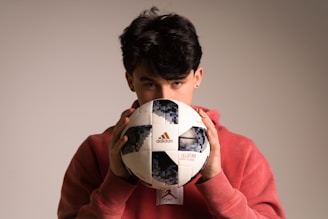 A person with dark hair partially obscured is holding a Telstar Adidas soccer ball close to their face while wearing a red hoodie. The background is neutral and softly lit, highlighting the person and the ball.