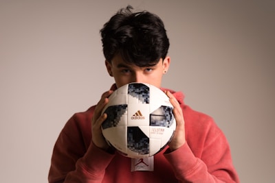 A person with dark hair partially obscured is holding a Telstar Adidas soccer ball close to their face while wearing a red hoodie. The background is neutral and softly lit, highlighting the person and the ball.