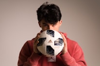 A person with dark hair partially obscured is holding a Telstar Adidas soccer ball close to their face while wearing a red hoodie. The background is neutral and softly lit, highlighting the person and the ball.