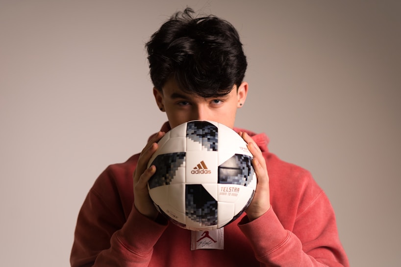 A person with dark hair partially obscured is holding a Telstar Adidas soccer ball close to their face while wearing a red hoodie. The background is neutral and softly lit, highlighting the person and the ball.