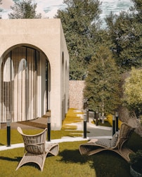 A modern architectural structure with large arching windows surrounded by a serene outdoor setting. The area features wicker chairs on a neatly trimmed lawn with stepping stones leading towards the building. Trees and shrubbery provide natural greenery, complementing the warm tones of the structure.